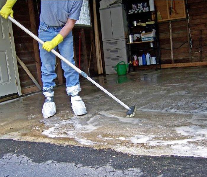 A person cleaning grease off a garage floor with soap and a scrub brush.