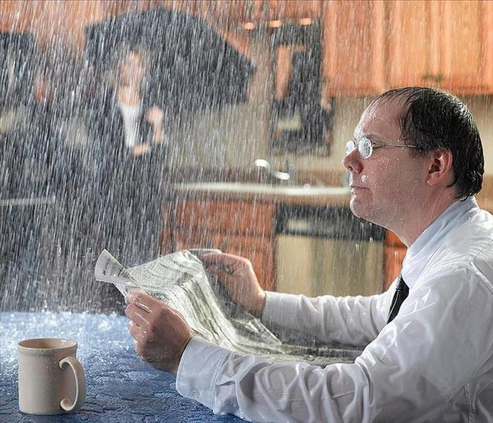 A man sitting in the kitchen at the table reading a paper with water pouring down on him.
